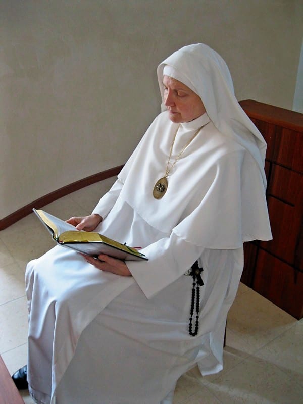 A sister in a white habit seated in a chair, reading from a large open book, a rosary visible at her side.