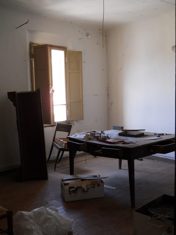A sparse, dusty room with peeling walls, an old table, a wooden chair and shuttered windows, awaiting renovation.