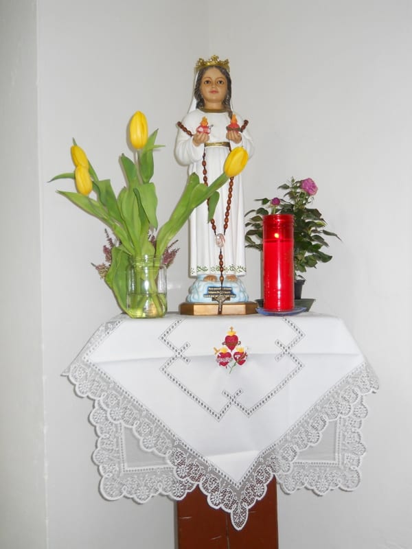 A small corner shrine with a statue of the Child Jesus dressed in white and gold, flanked by yellow tulips in a vase and a red vigil candle, on a white linen-covered shelf.