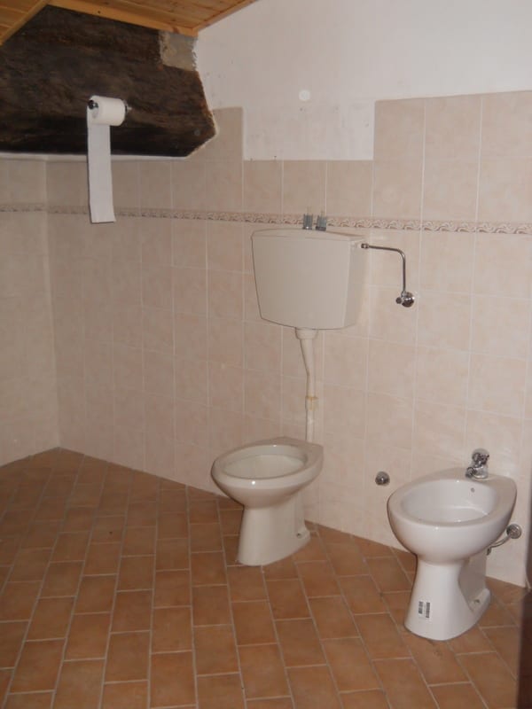 A tiled bathroom with a toilet and bidet installed but otherwise unfinished, with exposed rough masonry visible above the tile line.