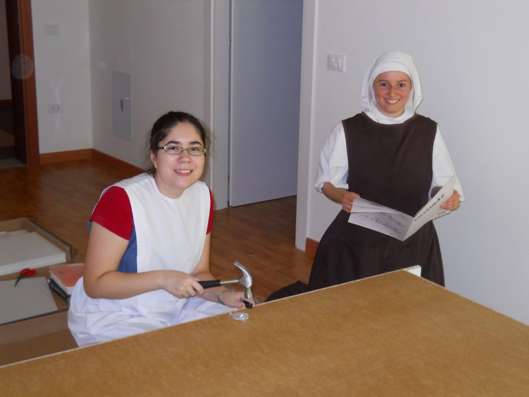 A young woman in lay clothes and a sister in a white habit smile at the camera across a wooden table in a bright, freshly finished room.