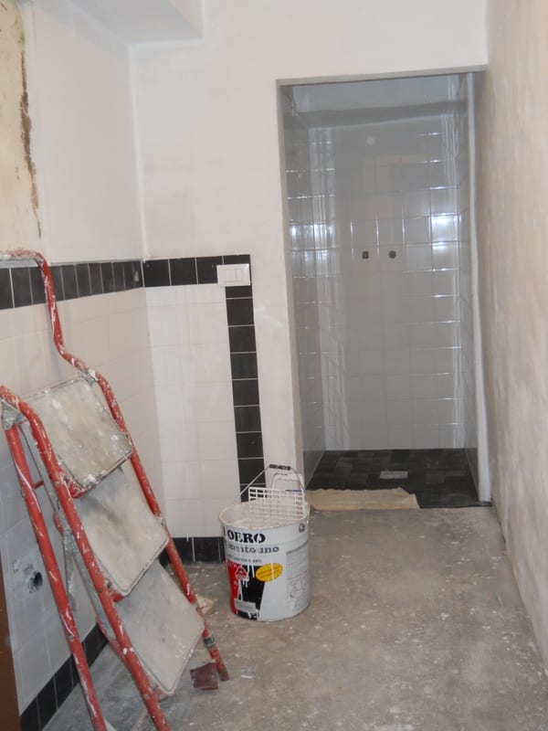A narrow corridor under renovation showing bare plaster walls, a new white door at the far end, and a bucket of materials on the floor.
