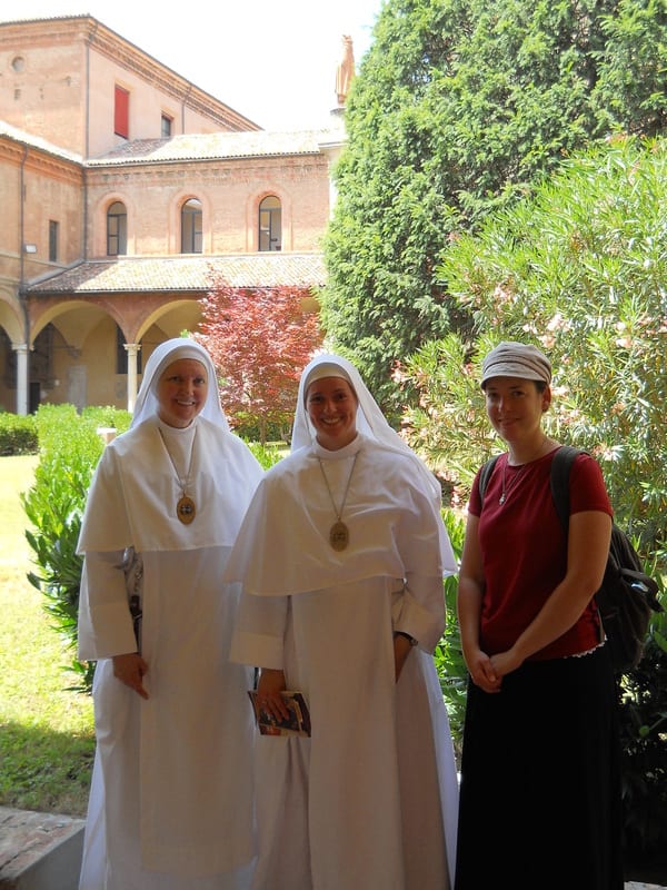 Two sisters in white habits and a woman in a sun hat pose together in a cloister garden with leafy trees behind them.