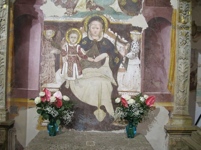 A painted altarpiece of the Madonna and Child flanked by two angels, decorated with fresh red and white flowers in vases.