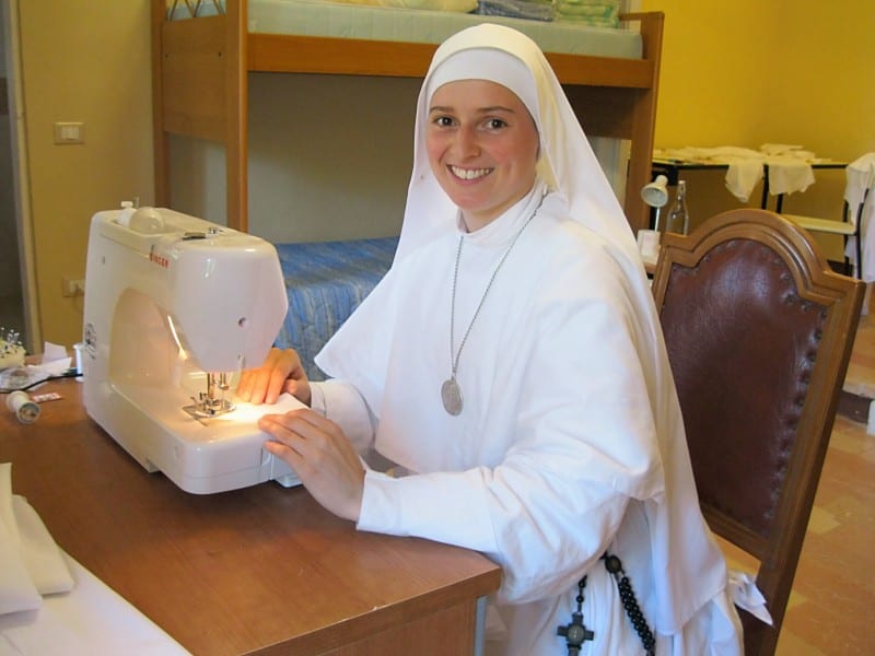 A smiling sister in a white habit works at a sewing machine, with fabric and a rosary visible on the table beside her.