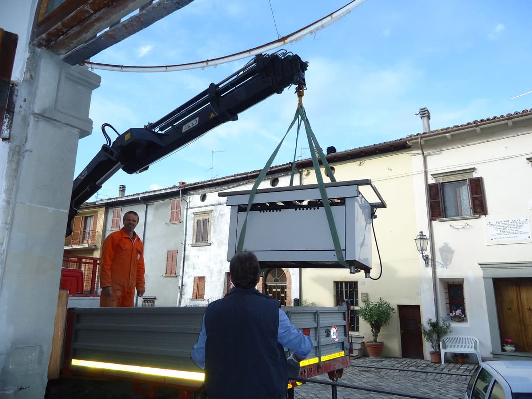 A truck-mounted crane lifts a large flat object over a narrow village street while two workers watch from below.