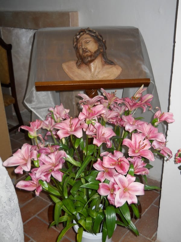 A large arrangement of pink lilies in front of a small wooden shrine holding a relief image of the crowned Christ.