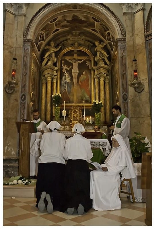 Several sisters in white habits and black veils kneel with heads bowed before an ornate golden altar during a liturgical ceremony.