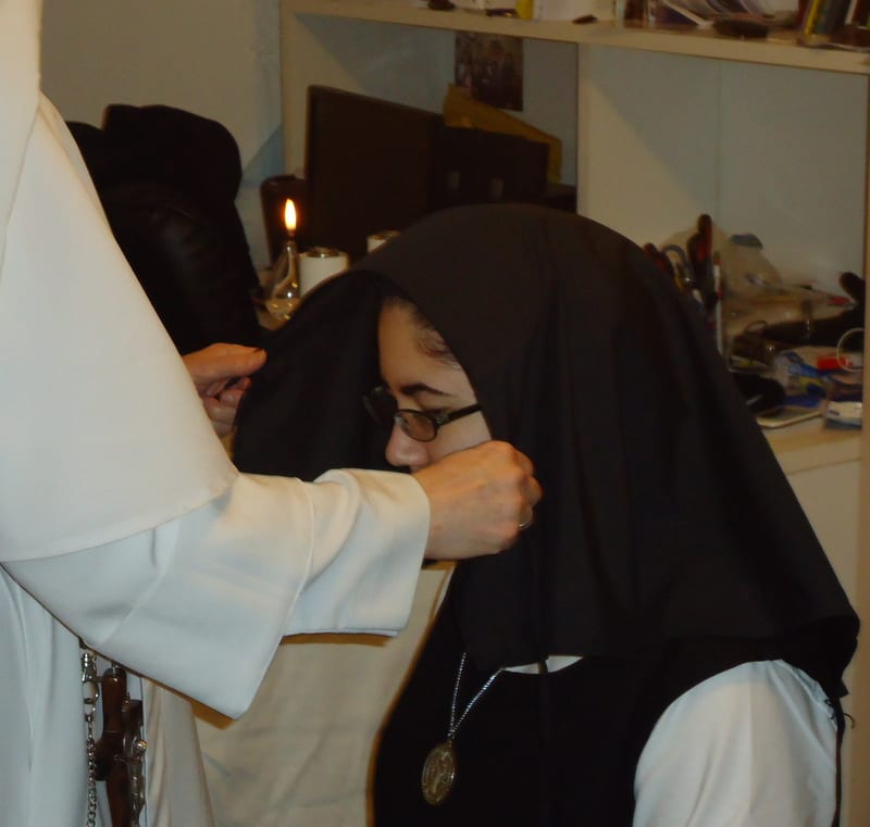 A sister in white adjusts and pins a black veil onto another sister who is bowing her head during the receiving of the veil.