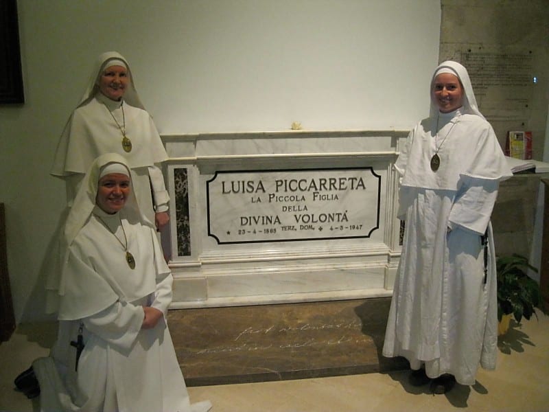 Three sisters in white habits flank the marble tomb of the Servant of God Luisa Piccarreta inscribed with her name and dates.