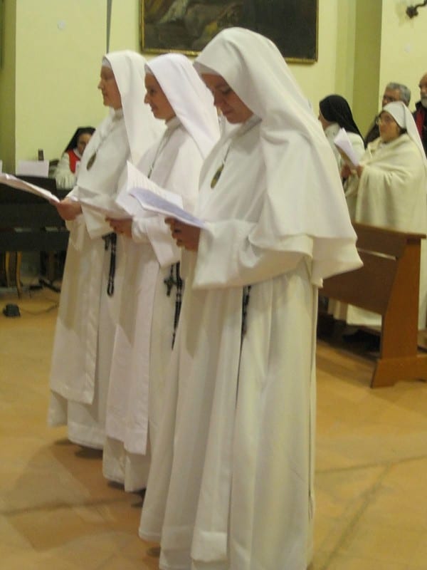 Three sisters in white habits and black veils stand in a row holding open booklets during a vows ceremony in a church.