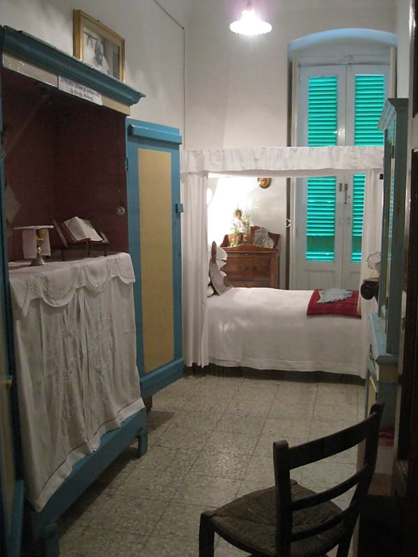 A simply furnished bedroom preserved as a historical room, with a white-draped bed, green shuttered window, wardrobe, and a chair.