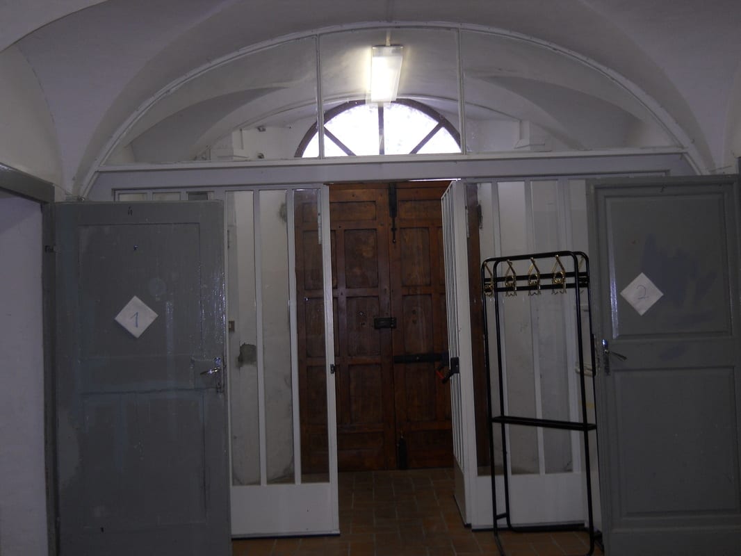 A dim vaulted entrance hall with grey metal doors on either side and a heavy wooden door straight ahead, lit by a single overhead light.