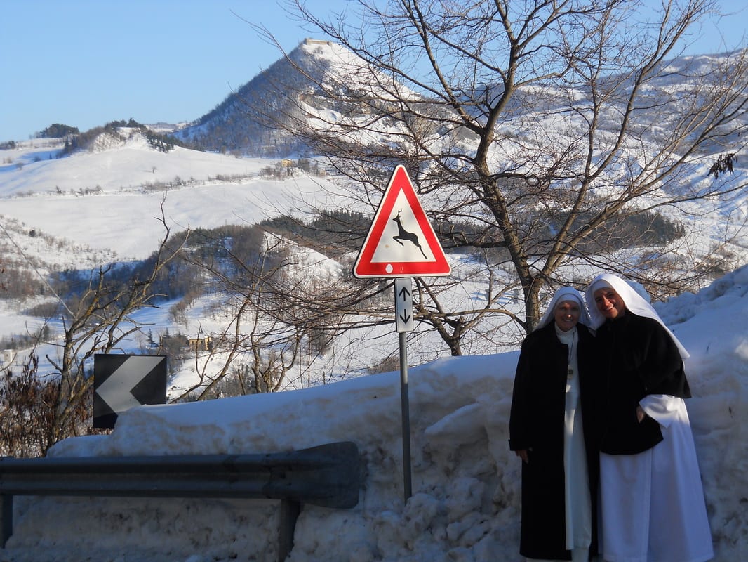 Two sisters in dark habits stand beside a deer-warning road sign, with snow-covered mountain ridges rising behind them.