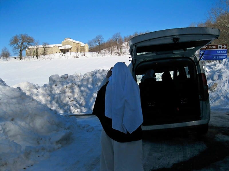 A sister in a dark habit stands at the open boot of a van in deep snow, with a snow-covered building visible in the background.