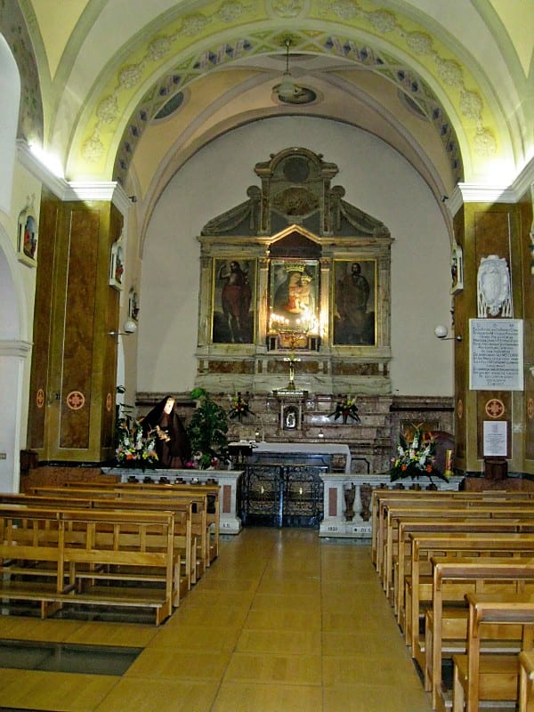 Interior of a small Italian church with wooden pews, frescoed arched ceiling, and an ornate gilded altarpiece at the far end.