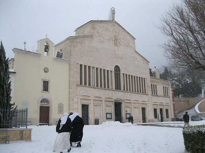 A large modern basilica with a stone facade dusted in snow, with a sister in a dark habit walking across the forecourt.