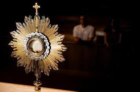 Gold monstrance holding the Blessed Sacrament, backlit against a dark interior, with a blurred figure visible behind it.