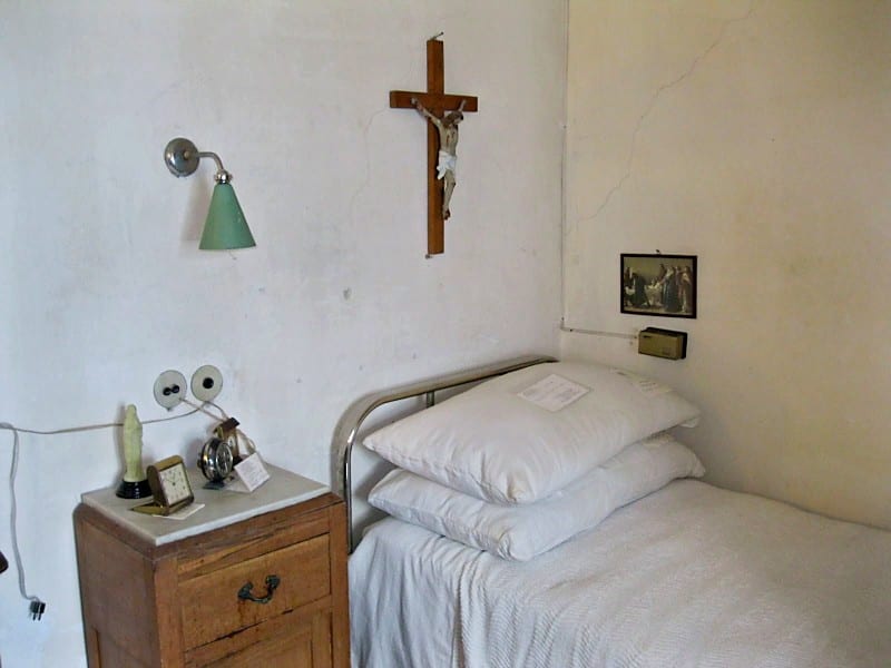 A narrow monastic cell with a metal-framed single bed, white pillow, a wooden nightstand with a lamp, and a crucifix on the wall above.