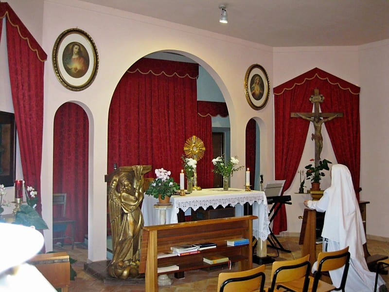 The interior of a small chapel with red curtained arches, a monstrance on the altar, a gold statue, and a veiled figure at prayer.
