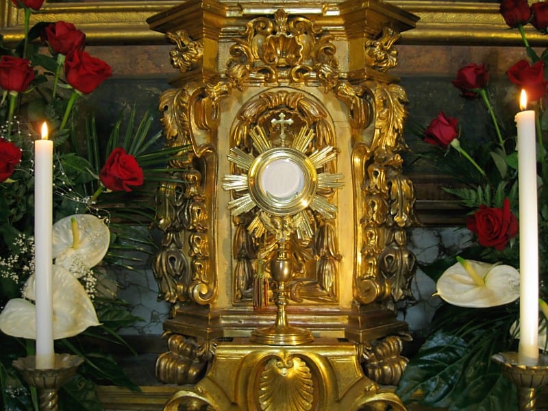 A close-up of an ornate gold monstrance surrounded by red roses and white flowers, flanked by two lit candles.