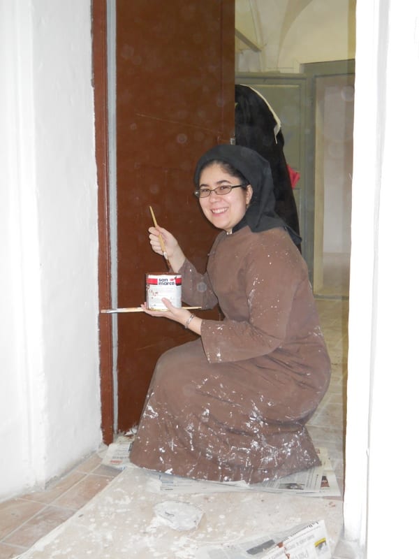 A sister in a brown work habit crouches on a drop-cloth and paints a door frame with a small brush, smiling at the camera.