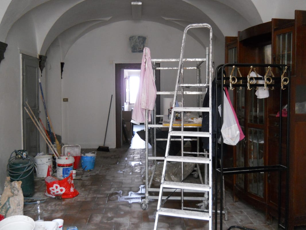 A vaulted room mid-renovation with a stepladder at centre, scattered paint buckets, tools, and a glass-fronted cabinet along one wall.