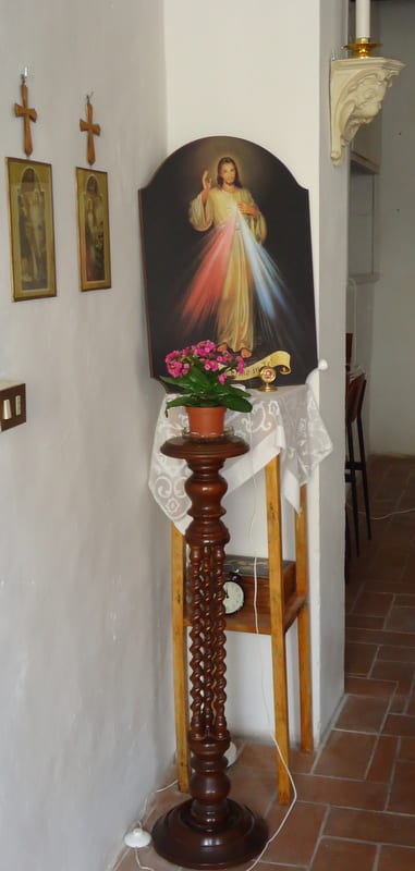 A corner of a corridor or room showing a Divine Mercy icon on a wooden stand, with a small cross visible on the wall beside it.