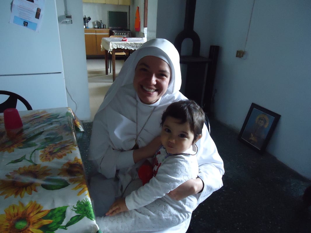 A sister in a white habit hugs a small toddler in a domestic kitchen, with a sunflower-print tablecloth and a wood-burning stove visible.