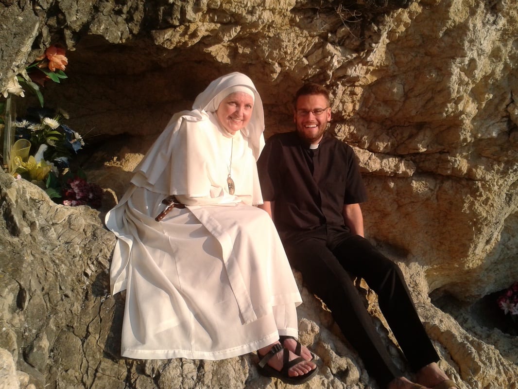 A sister in a white habit and a young man in clerical black seated together in the hollow of a rocky grotto, smiling, with flowers placed nearby.