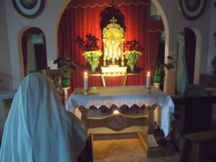 A sister in a white habit kneeling before a lit altar with a gold monstrance at its center, two candles, and red fabric draping behind, in a small chapel interior.