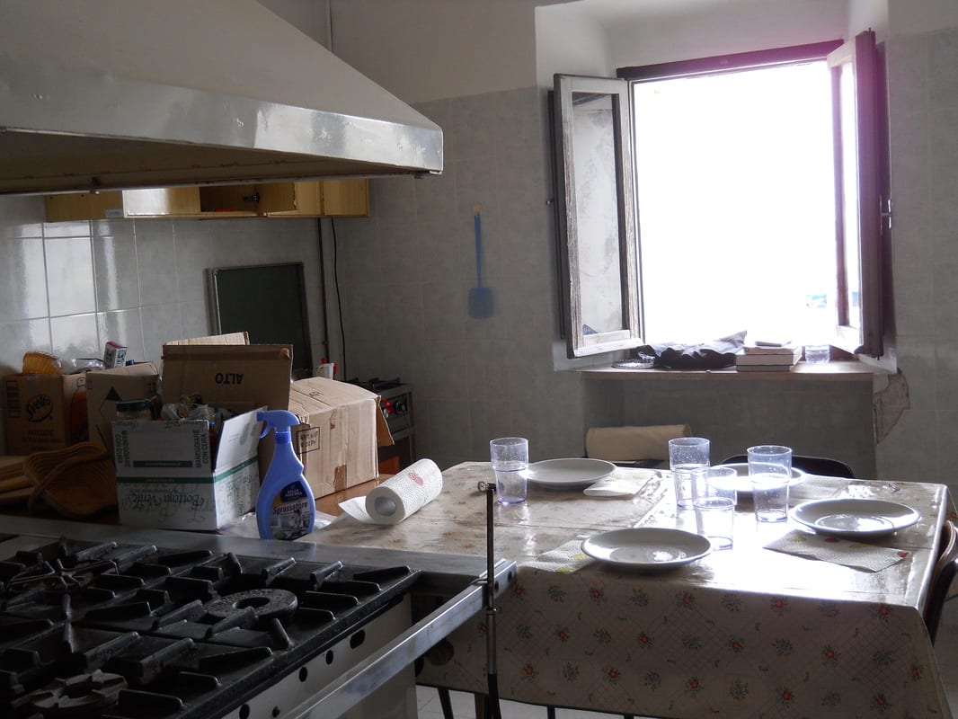 A dated kitchen with a gas hob, large extractor hood, a table set with plates and glasses, and cardboard boxes stacked near the window.