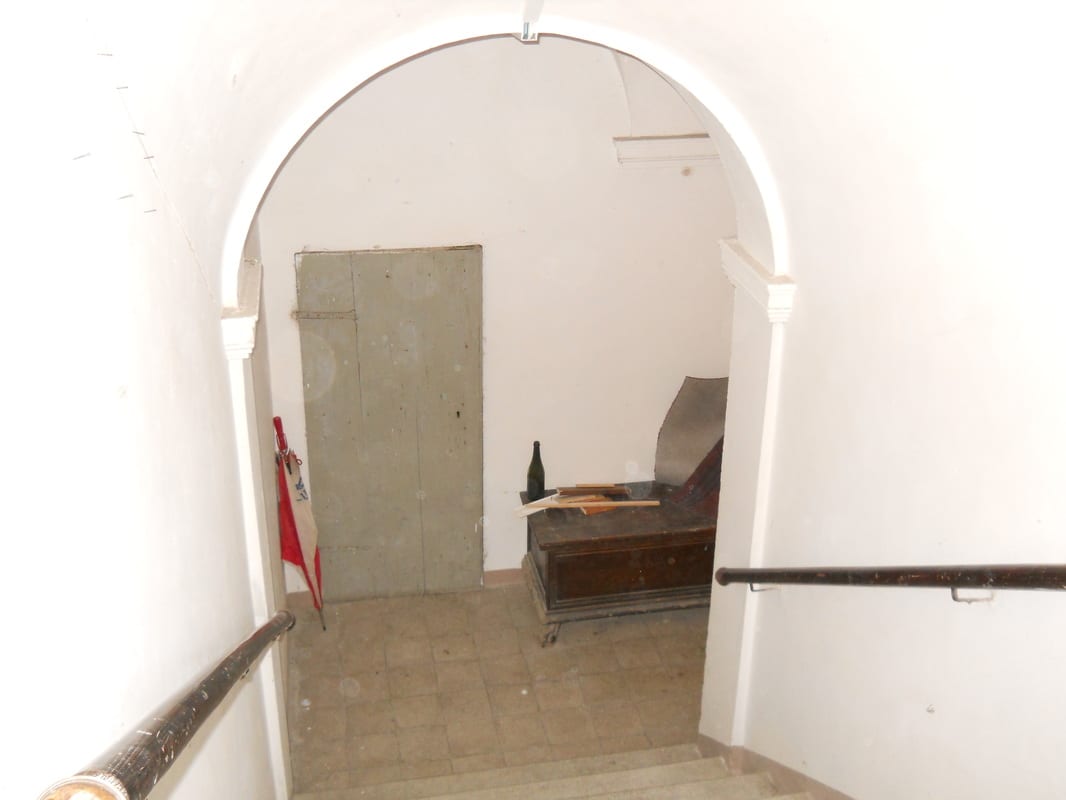 View through a round arch up a bare stairwell landing, with rough plaster walls and a metal handrail, looking toward a doorway.