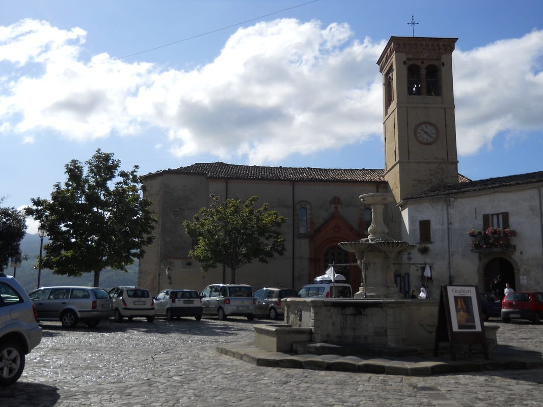 The main piazza of a small Italian hill town with a stone clock tower, a church, parked cars, and a central fountain.