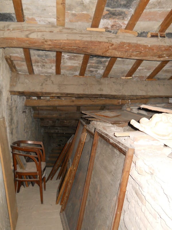 An unfinished attic or loft space with exposed timber ceiling joists, bare stone walls, and old wooden boards and debris on the floor.