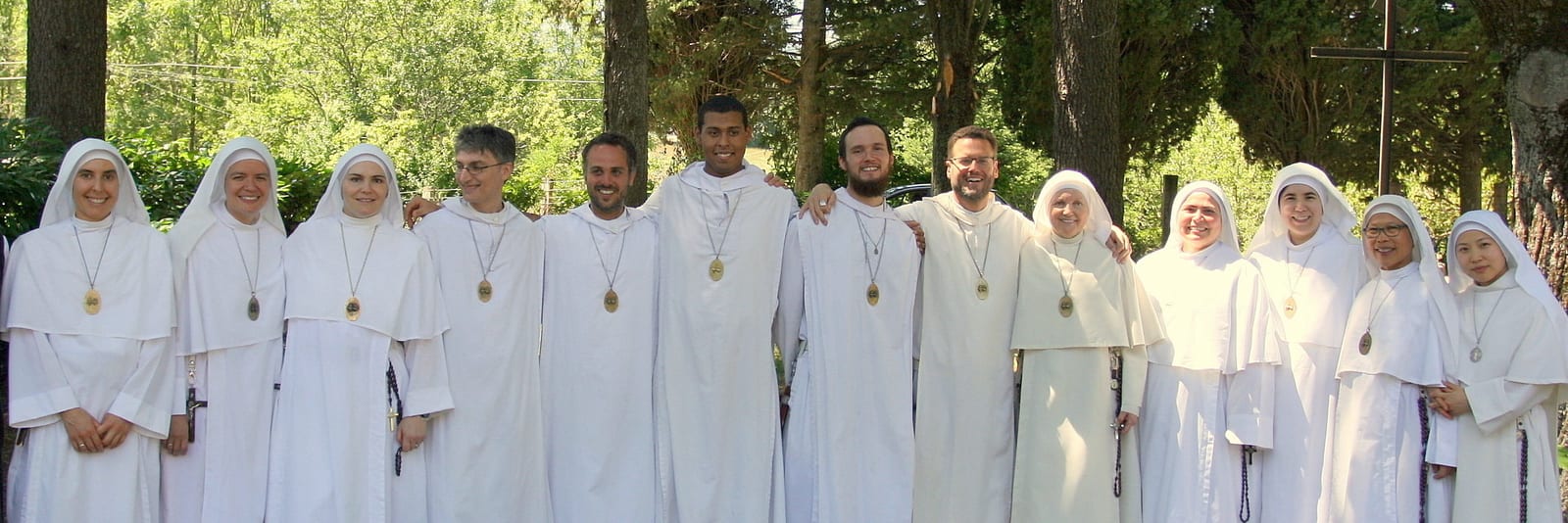 The community standing in a long line outdoors among trees: sisters in white habits and veils and brothers in white robes, each wearing a medal.