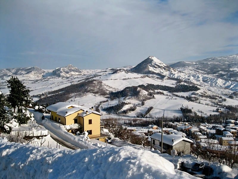 A yellow house and neighbouring buildings seen from above in a snow-covered hillside village, with a rocky peak beyond.