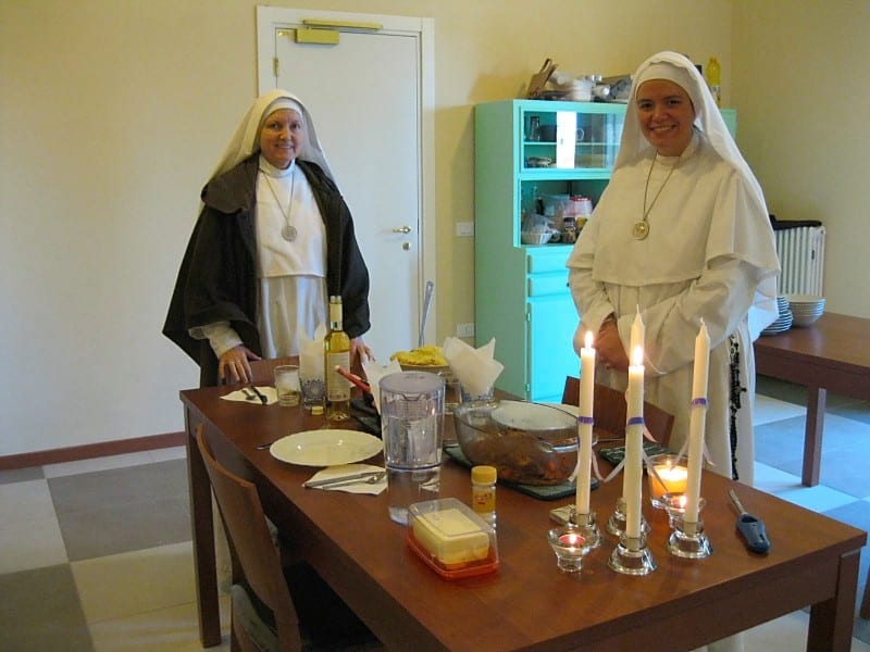 Two sisters in black and white habits stand on either side of a wooden dining table set with candles, glasses, and food for a meal.