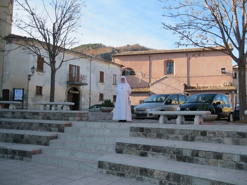 A sister in a white habit stands on broad stone steps in a piazza, with a hillside village and bare trees behind her.