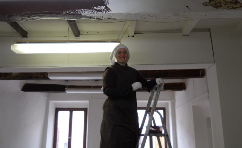 A sister in a black habit and white veil stands on a stepladder, working on a damaged ceiling during renovation.