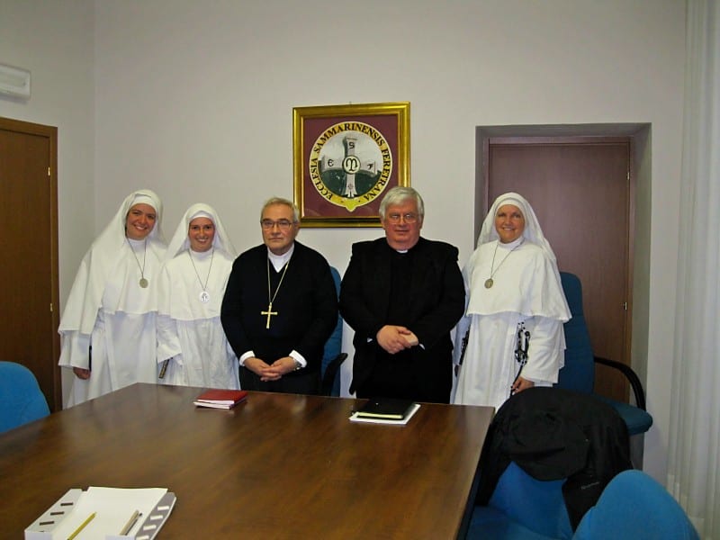 Three sisters in white habits, a bishop in purple, and a vicar general pose around a conference table in an office with a diocesan seal on the wall.