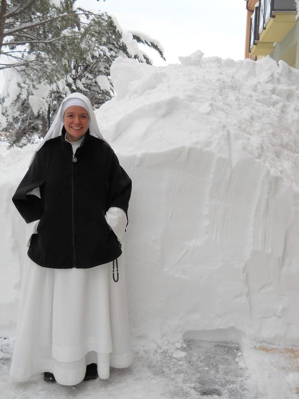 A sister in a black coat and white habit stands smiling beside a large mound of snow nearly as tall as she is.