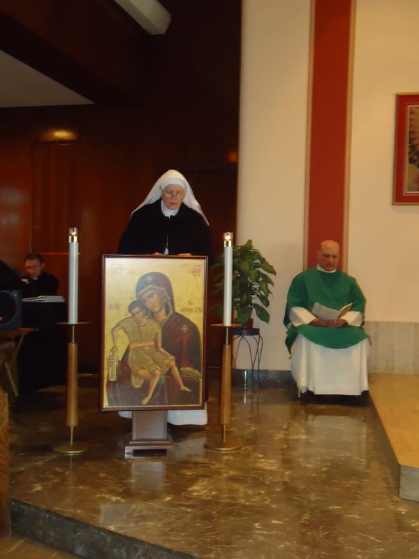 A sister at a lectern and a priest in green vestments at the altar during a vows ceremony, with an icon of Mary and Child displayed prominently.