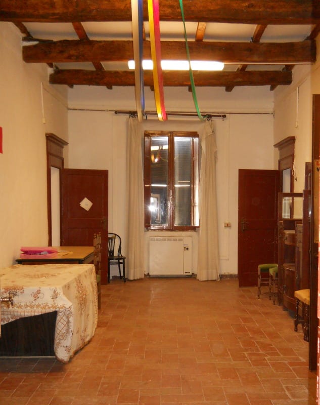 A bare, tile-floored room with wooden-beam ceiling, colourful streamers hanging from a light fixture, and sparse old furniture.