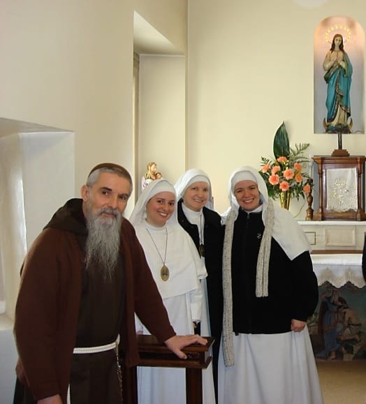A Capuchin friar and three sisters in white habits stand before a small altar with a statue of Our Lady and floral arrangements.