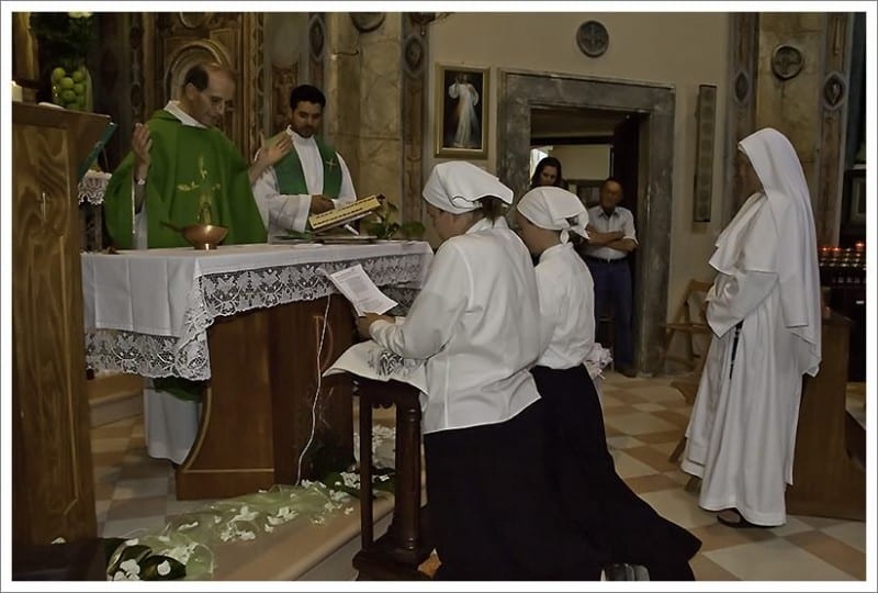 Two candidates in white veils kneel before a priest at the altar during an investment ceremony, with a sister standing to the right.