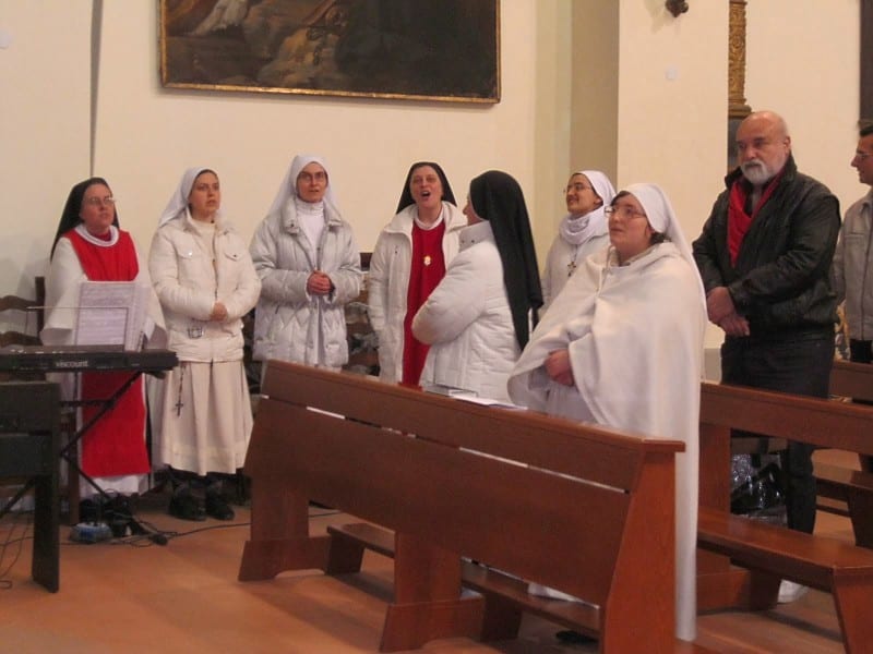 A group of sisters in white and red habits singing together in a church nave, with a man standing to the right.