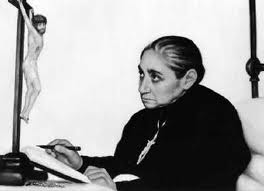 Black-and-white photograph of the Servant of God Luisa Piccarreta at a desk, pen in hand, with a crucifix beside her.