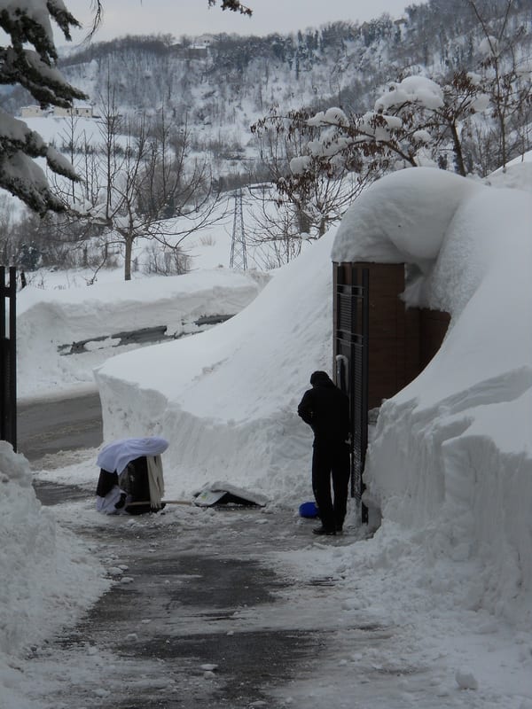 Two figures shovelling a narrow snow-bound path between high snow walls outside a building, with a hillside visible beyond.