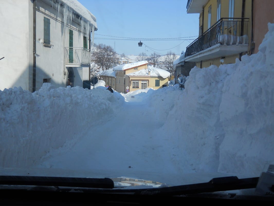 A view from inside a car looking down a narrow village street with walls of ploughed snow towering on both sides.